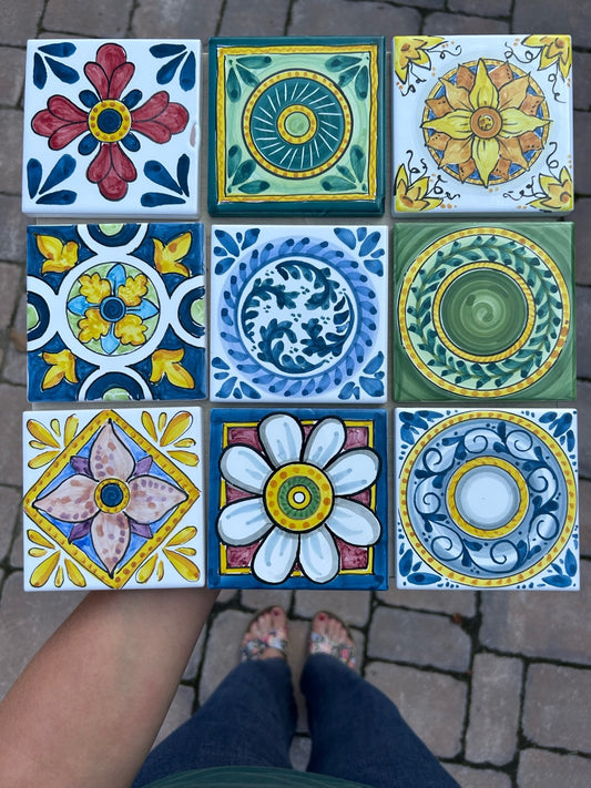 Nove mattonelle quadrate accostate a griglia su un pavimento di pietra: rosoni, margherite e girandole nei toni del blu, del giallo e del verde.