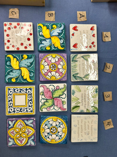 Dodici mattonelle disposte a griglia su un telo blu: decori geometrici, fiori e frasi.