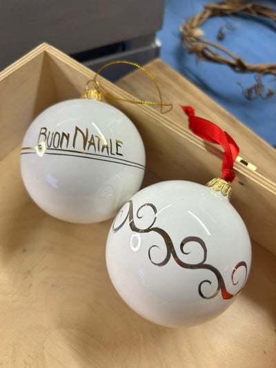 Due sfere bianche in una cassetta di legno: una con «Buon Natale», una con un ricciolo dorato e il nastro rosso.