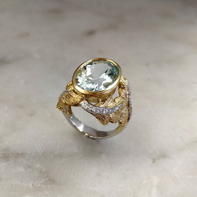 Three-quarter view on marble: the oval aquamarine in the yellow gold bezel, the engraved leaves and the ribbon of diamonds