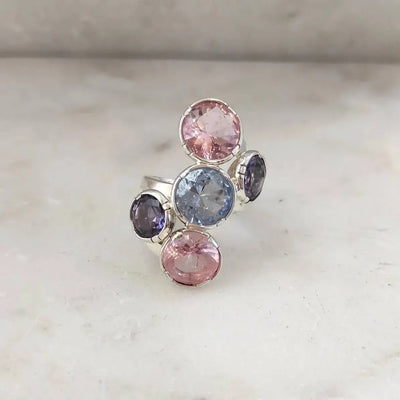 L'anello posato su marmo chiaro, visto dall'alto: le tre paste vitree tonde in fila obliqua e le due ovali viola incastrate ai lati.
