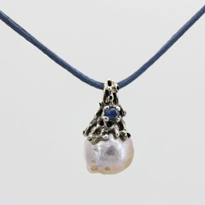 The pendant seen from the front on the blue silk cord: the burnished silver cap, the sapphire and the white baroque pearl