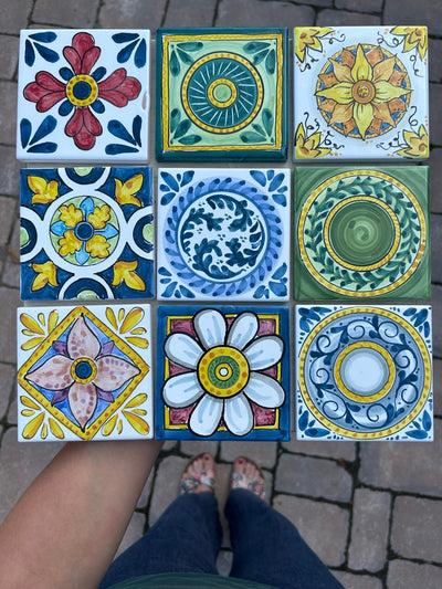 Nove mattonelle quadrate accostate a griglia su un pavimento di pietra: rosoni, margherite e girandole nei toni del blu, del giallo e del verde.