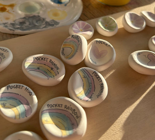 Molti sassi bianchi con arcobaleni dipinti e la scritta «pocket rainbow», allineati su un vassoio di legno.