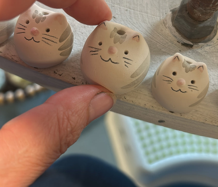 Tre gattini di ceramica allineati su una mensola, uno tenuto fra le dita.