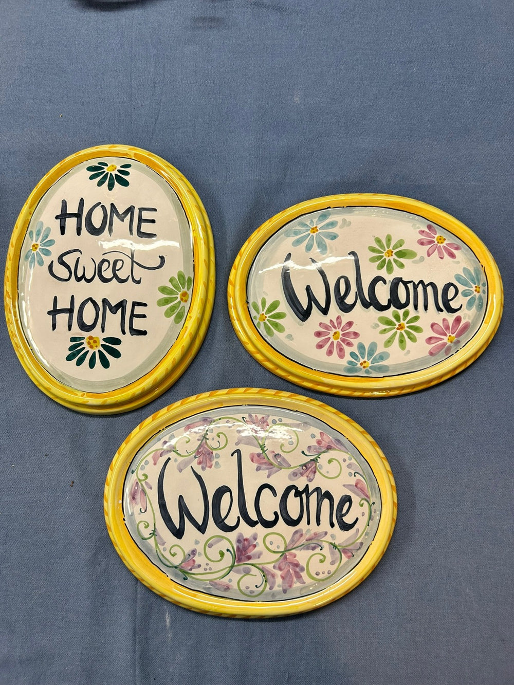 Tre targhe ovali su un telo blu, con «Home sweet home» e «Welcome» e il bordo giallo a treccia.