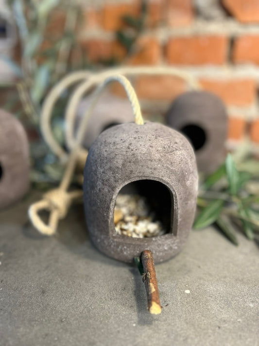 La mangiatoia di terracotta appoggiata a terra davanti a un muro di mattoni, con il rametto che fa da posatoio e i semi dentro.