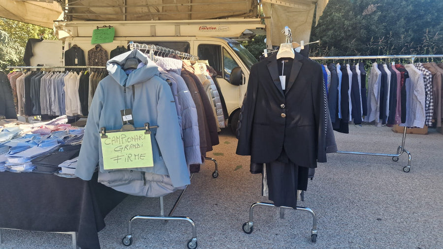 Il banco di Alessandro Benini: in primo piano un piumino col cartello «campionari grandi firme» e un blazer nero, dietro le file di camicie e il furgone
