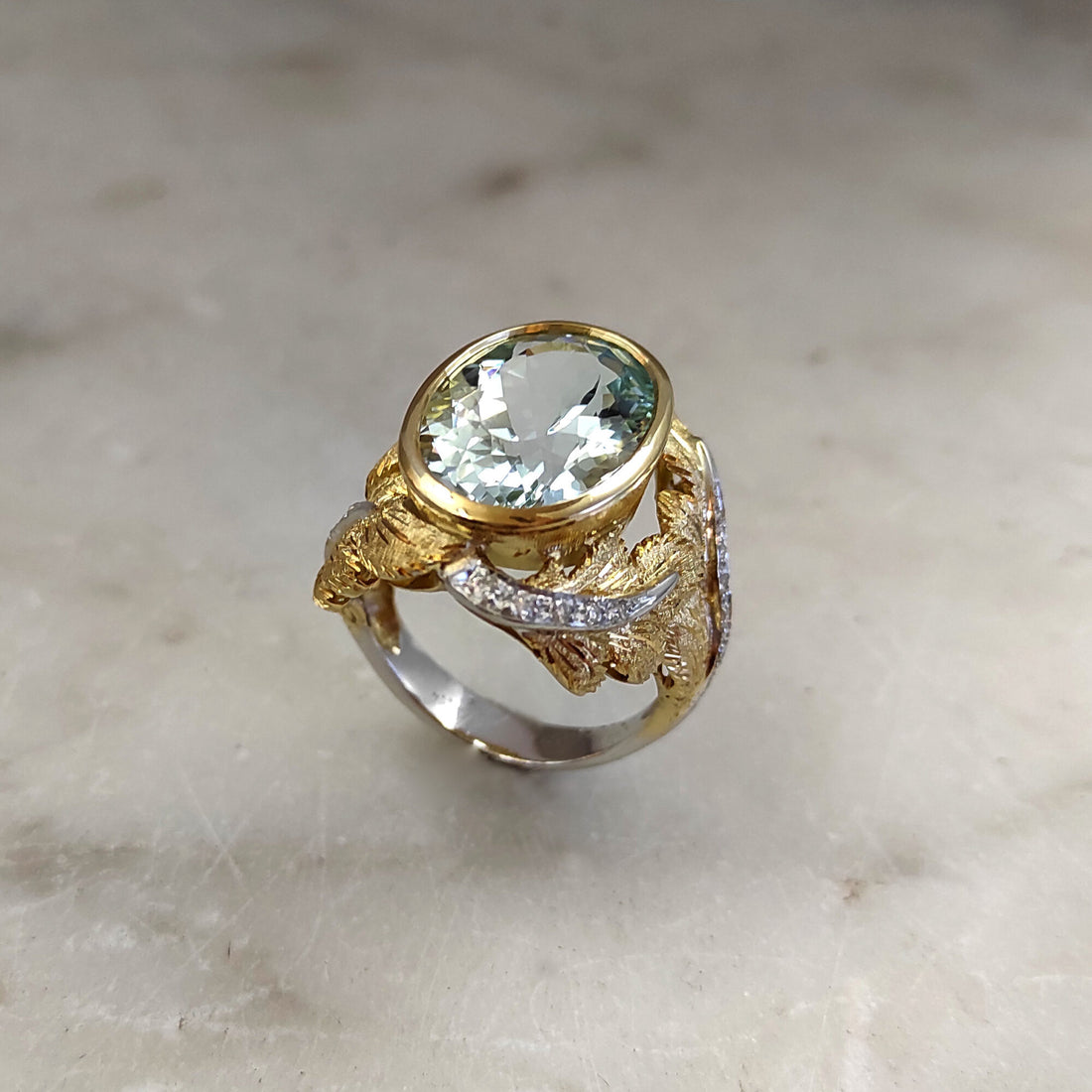 Three-quarter view on marble: the oval aquamarine in the yellow gold bezel, the engraved leaves and the ribbon of diamonds