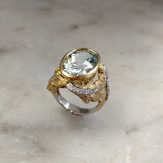 Veduta di tre quarti su marmo: l'acquamarina ovale nel castone d'oro giallo, le foglie incise e il nastro di diamanti