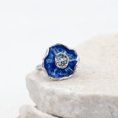 L'anello su una lastra di pietra chiara, la corolla vista di fronte con lo smalto blu e il topazio al centro
