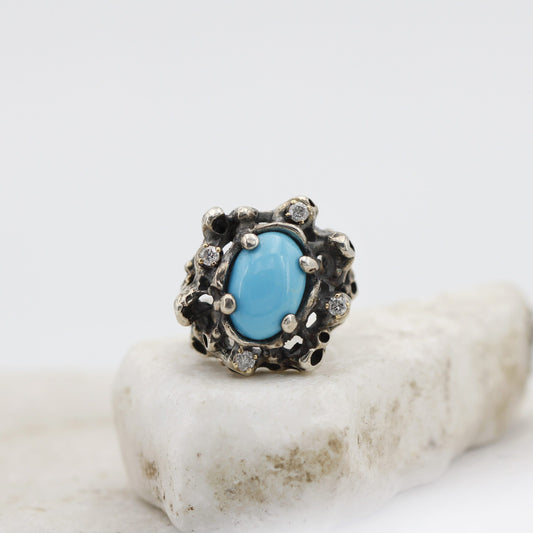 Il turchese a cabochon ovale al centro dell'argento brunito e bucato, con i quattro diamanti grigi nei castoni d'oro