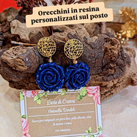 Orecchini con rosa di resina blu e perno dorato traforato, su un ceppo di legno, con il cartello «orecchini in resina personalizzati sul posto»