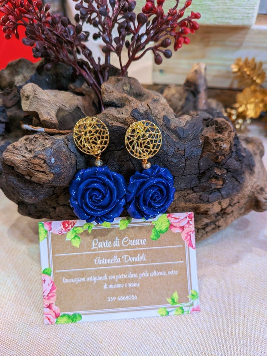 Gli stessi orecchini con la rosa blu accanto al cartellino della bottega, su un ceppo di legno