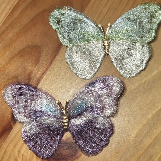 Due spille a farfalla ricamate su tulle, una verde e argento e una viola e grigia, posate sul legno