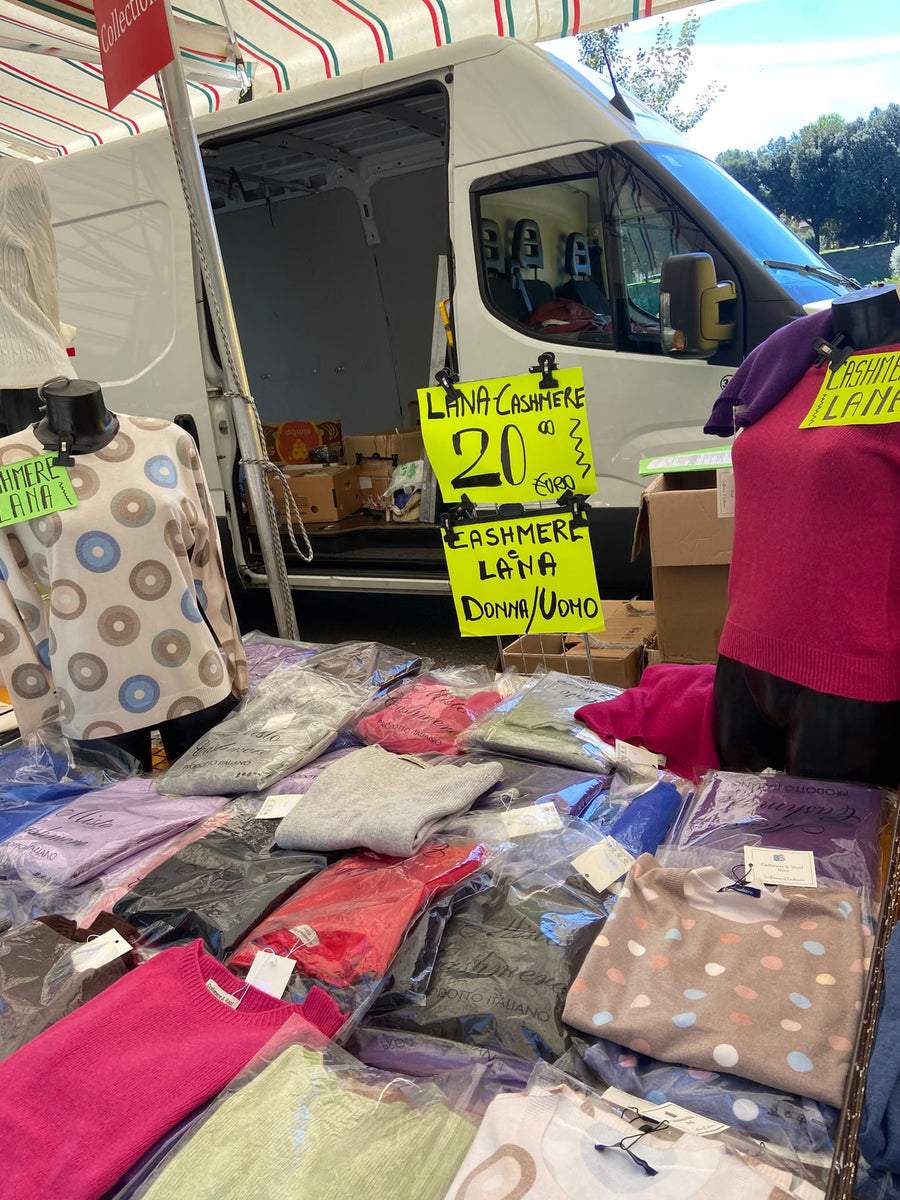 Il banco di maglieria Castellani con i cartelli gialli del cashmere e della lana, maglioni impilati e il furgone aperto dietro