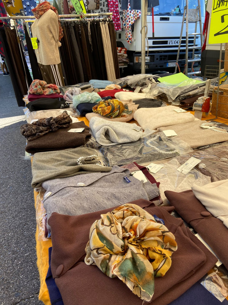 Il banco visto di lato: pile di maglioni nei toni del marrone e del beige, foulard e un cappotto cammello appeso