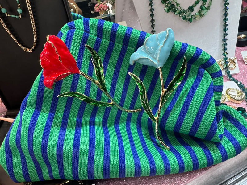 Due spille smaltate a fiore, una rossa e una azzurra, appuntate su una borsa a righe verdi e blu