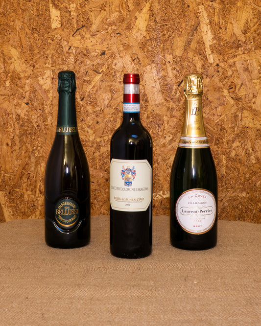 Le 3 bottiglie della selezione «Cenone di Capodanno» in piedi davanti al fondo del set: Valdobbiadene Superiore Extra Dry Bellussì, Rosso di Montalcino Ciacci Piccolomini e Champagne Laurent Perrier Brut.