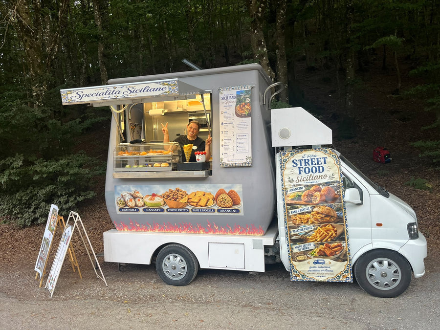 The Le Delizie di Emy truck open in the woods, with the “Specialità siciliane” awning, Erminda at the hatch and the menu hanging on the side
