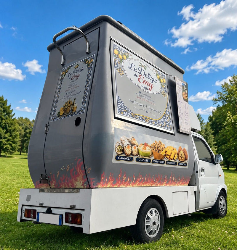The truck seen from behind, on a lawn: the sign “Le Delizie di Emy, friggitoria rosticceria pasticceria siciliana”, the painted flames and the strip with cannoli, cassate, fried cone, pane e panelle, arancini