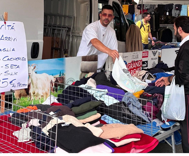 Luca Carlesi al banco mentre serve un cliente, fra le pile di maglioni e un cartello scritto a mano con i nuovi arrivi