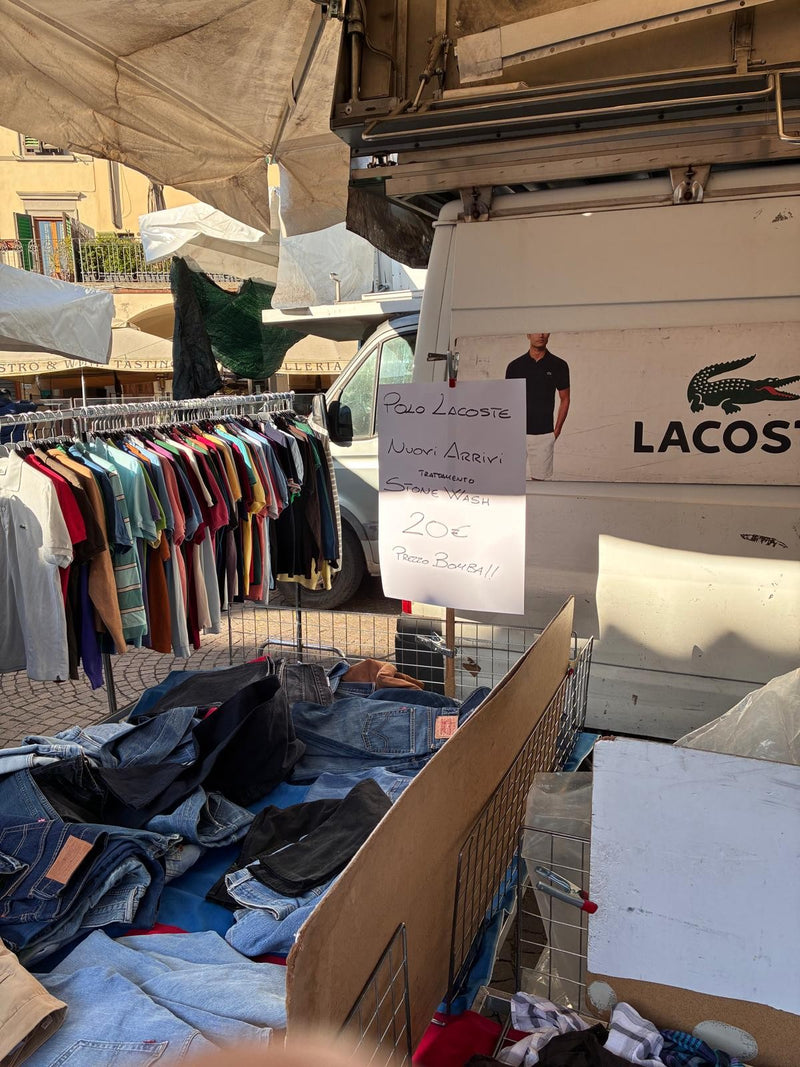 Polo appese e jeans ammucchiati in una cassa, con un cartello scritto a mano e il furgone del banco