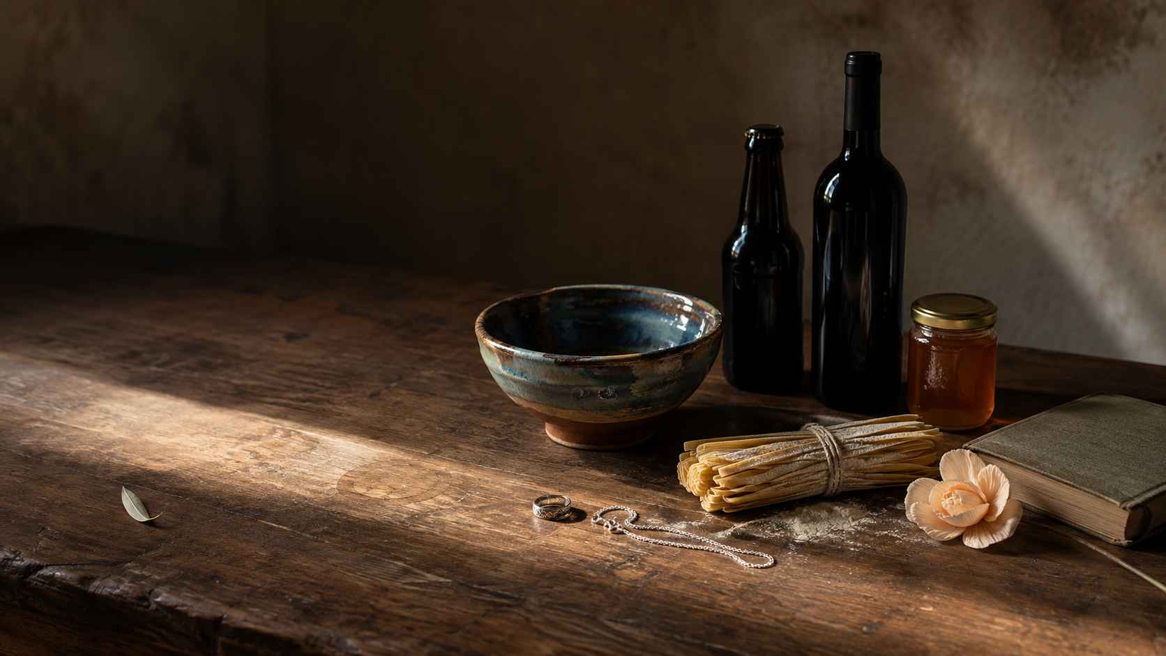 Un tavolo di noce con sopra la merce di più mestieri toscani: una scodella di ceramica smaltata, una bottiglia di birra e una di vino, un vasetto di miele, un mazzo di pasta secca, un anello e una catenina d'argento posati sul legno, un fiore di carta e un libro
